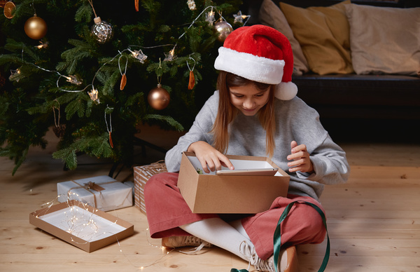 A little girl in a Santa hat sitting on the floor under a Christmas tree enthusiastically examining Christmas gifts in an open box with a green ribbon and a garland nearby