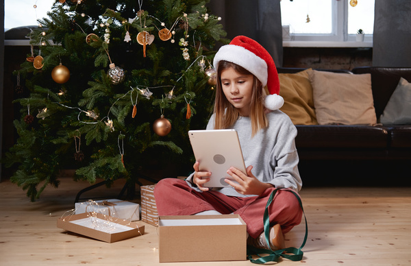 A girl with brown loose hair in a Santa hat sitting on the floor with a box a ribbon and a garland around under a decorated Christmas tree taking a tablet out of a gift box looks at it