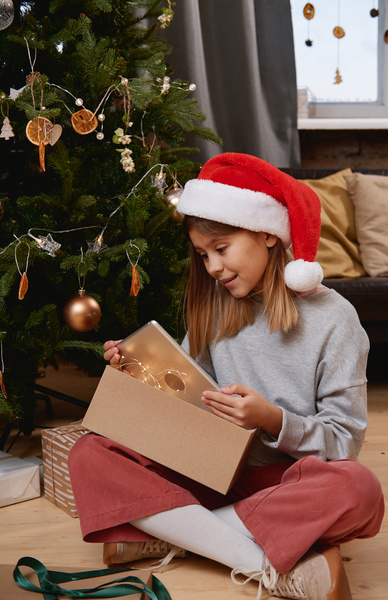 A girl with brown loose hair in a Santa hat sitting on the floor under a decorated Christmas tree takes out a tablet from a gift box that was decorated with a green ribbon and a garland