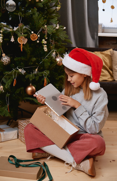 A little girl in a Santa hat looking at a tablet that she took out of a gift box as a Christmas present while sitting under the Christmas tree