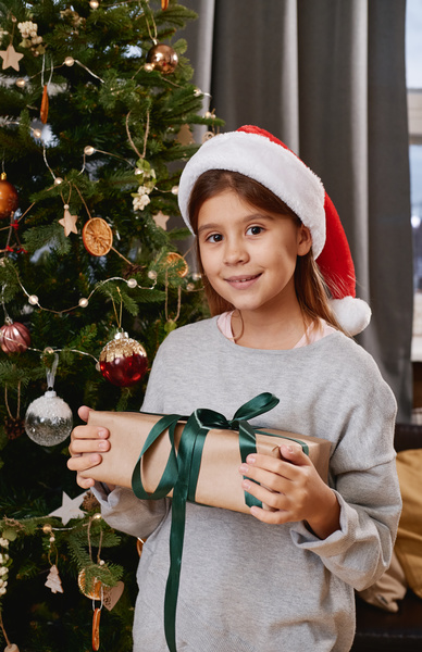 A little girl with loose hair in a Santa hat stands at a decorated Christmas tree with a gift in a craft wrapper with a green ribbon and smiles