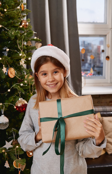 A girl in a Santa hat stands against the background of a decorated Christmas tree and with an admiring look holds a Christmas present in a craft wrapper with a green ribbon in her hands