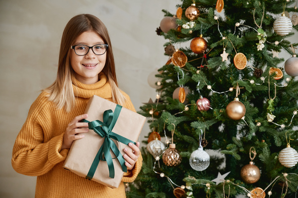 A girl in a yellow sweater and glasses standing next to a decorated Christmas tree holding a Christmas present in a craft package with a green ribbon