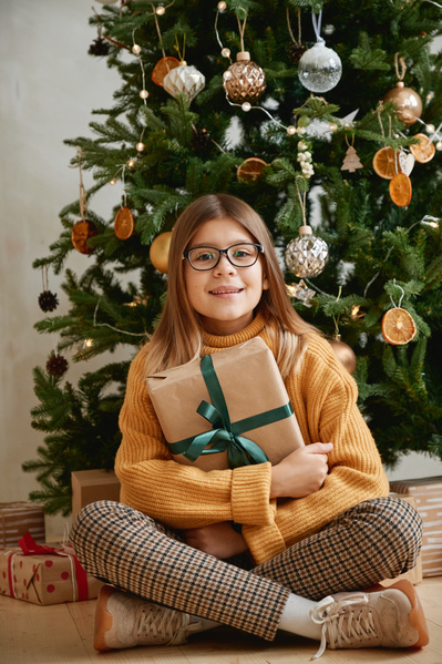 A girl in a yellow sweater plaid pants and glasses sitting in a lotus position on the floor under a decorated Christmas tree hugging a Christmas gift in kraft paper decorated with a green ribbon