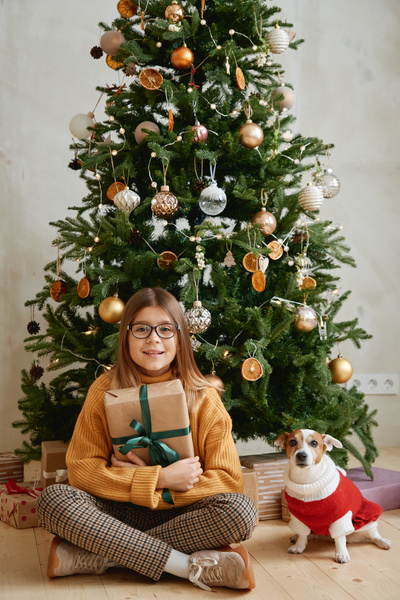 A girl in a yellow sweater checkered pants and glasses sitting in a lotus position on the floor under a decorated Christmas tree with a Christmas gift in kraft paper with a green ribbon and a dog in a red suit
