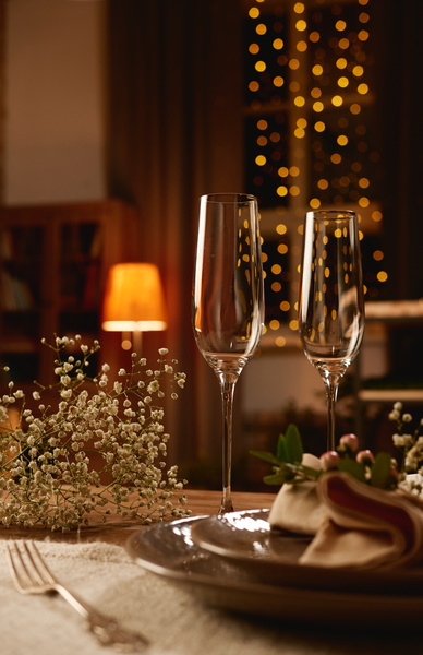 Champagne glasses stand on a laid table decorated with flowers in a cozy room with dim lights and a Christmas atmosphere