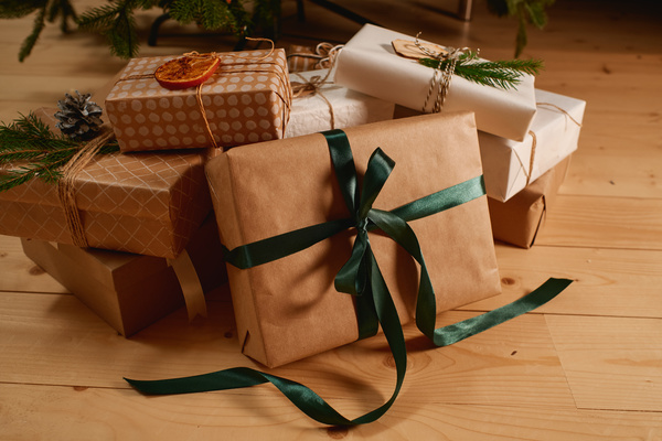 A gift box wrapped in craft paper tied with a green ribbon stands next to other gifts that are decorated with fir twigs and cones and dried slices of orange