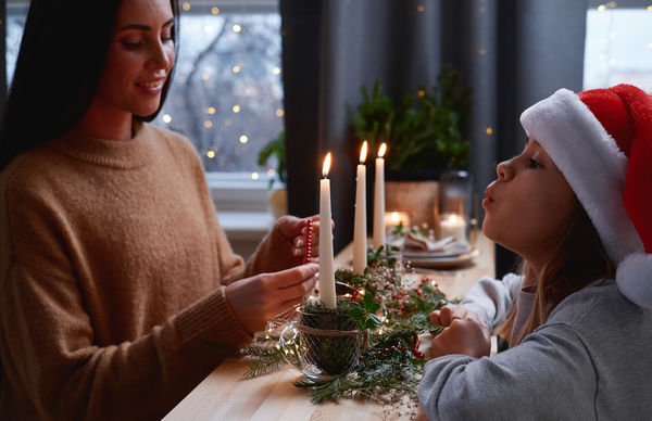 A woman with dark hair makes a tabletop Christmas decoration and her daughter in a Santa hat blowing out a candle