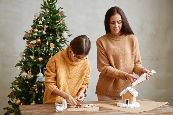Mom decorates a gingerbread house with cream from a pastry bag while her daughter rolled up her sleeves adds sprinkles on gingerbreads of different shapes standing at the table against the background of a Christmas tree