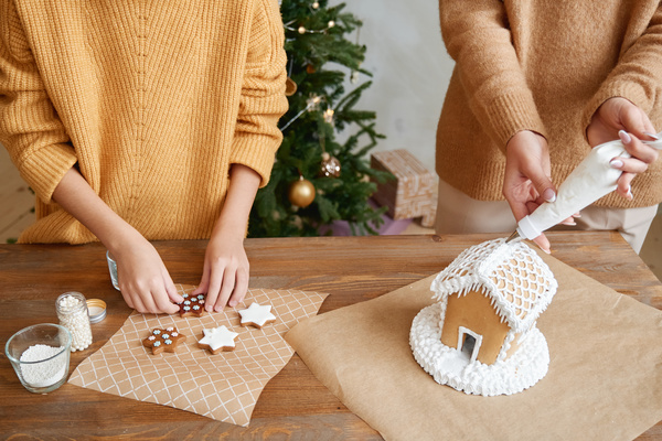 The daughter with the sleeves her sweater rolled and adds a pastry sprinkle to the gingerbread and her mother creams the gingerbread house using a pastry bag