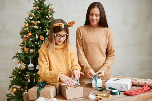 A mother in a beige sweater ties a ribbon on a gift while her daughter in a Christmas headband puts a Christmas bauble in a box standing at a table on which packaging materials are laid out