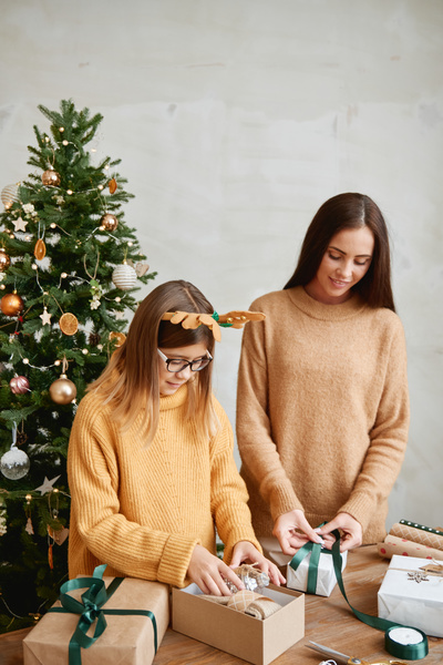 Mom and her daughter in sweaters enthusiastically pack gifts for their friends at Christmas standing at a table on which there are gifts ribbons and rolls of wrapping paper and there a Christmas tree behind
