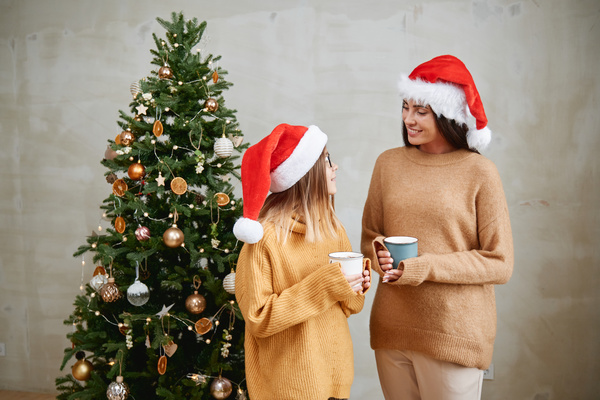 Mom and daughter in yellow sweaters and Santa hats looking at each other hold white and blue mugs with cocoa on the background of a Christmas tree