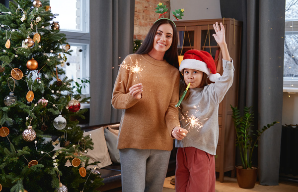 A mother in a sweater and a Christmas headband with burning sparklers in her hands stands with her daughter in a Santa hat enthusiastically blowing a festive whistle in the living room next to the Christmas tree
