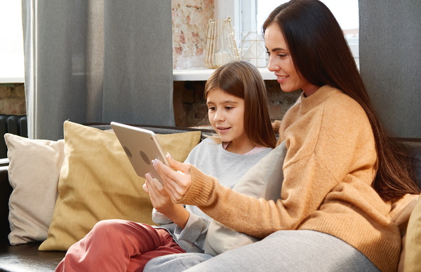 Mom with dark long hair dressed in a sweater with her daughter in a gray sweatshirt sitting on the couch with pillows with a tablet in their hands watch cartoons
