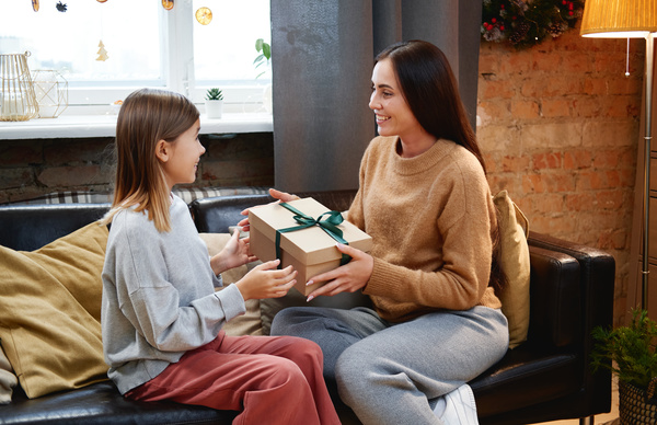 A woman with dark long hair in a sweater passes her admiring daughter a box decorated with a green bow with a Christmas gift