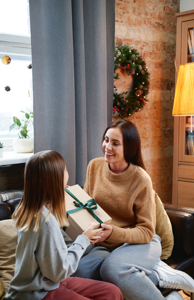 A mother in a sand-colored sweater smiling gives her daughter a Christmas gift in a box decorated with a green bow sitting on the sofa in the living room decorated for Christmas
