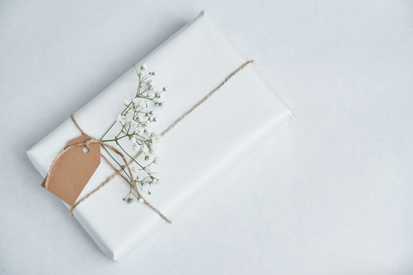 A gift in a white matte wrapper a white flower decorating it tied and with a thin rope with a tag on it