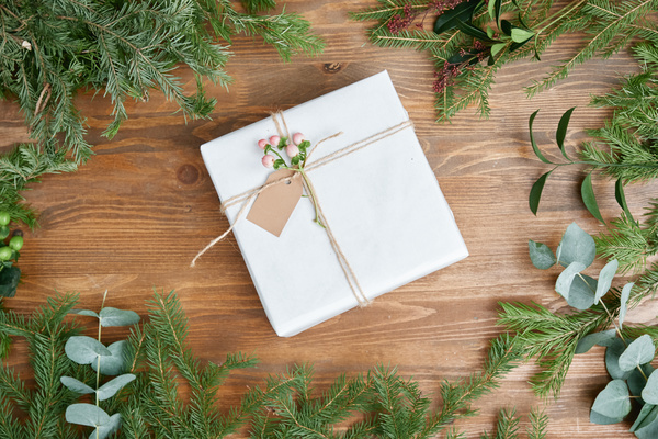 A light gift box tied with twine decorated with a pink flower and a note on the table among Christmas tree branches and other plants