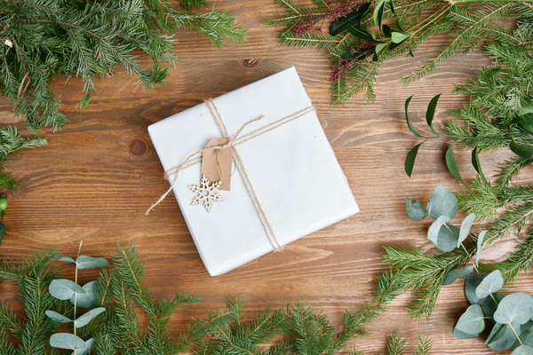 A light present box tied with twine decorated with a wooden snowflake and a note on the table among Christmas tree branches and other plants