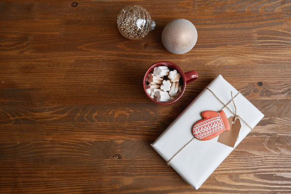 A light gift box tied with string with a note and a gingerbread mitten in icing on it on the table with a mug of cocoa with marshmallows and Christmas baubles in sequins