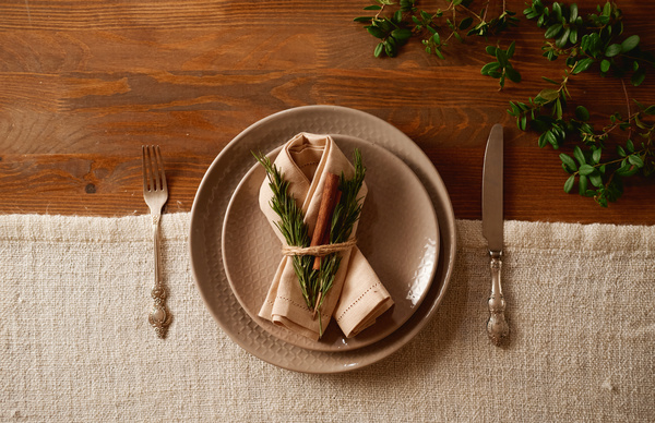 Plates with a napkin on them decorated with thuja twigs and a cinnamon stick for Christmas dinner on a table with cutlery and a green plant