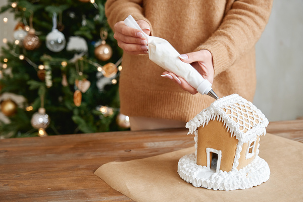 A woman in a sand-colored sweater decorates a gingerbread house with white cream from a pastry bag standing at a table against the background of a Christmas tree