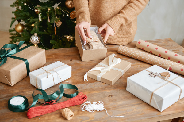 A woman in a beige sweater puts a knitted scarf in a box as a Christmas gift standing at a table on which there are gifts and packaging materials