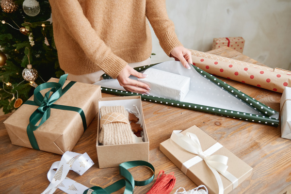A woman in a sand-colored sweater wraps a Christmas present box in green polka dot paper while standing at a table with ribbons and gift boxes on it