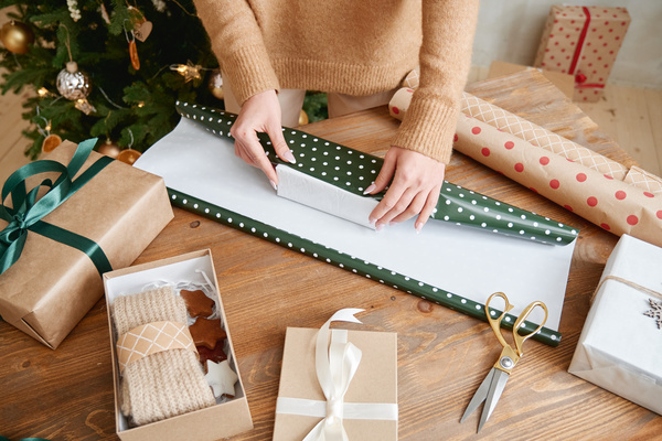 A woman in a beige sweater covers a box with a Christmas present with green polka dot paper standing at a table on which there are boxes and packaging materials