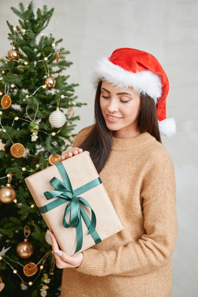 A woman with dark hair in a Santa hat and a beige sweater holds a Christmas gift wrapped in kraft paper and with a green ribbon bow and looks at it standing against the background of the Christmas tree