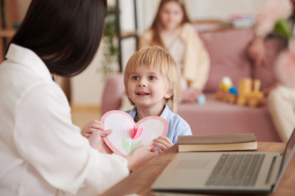 A small blond boy presents his mother sitting at a desk with a laptop a homemade postcard in the form of a heart with a flower