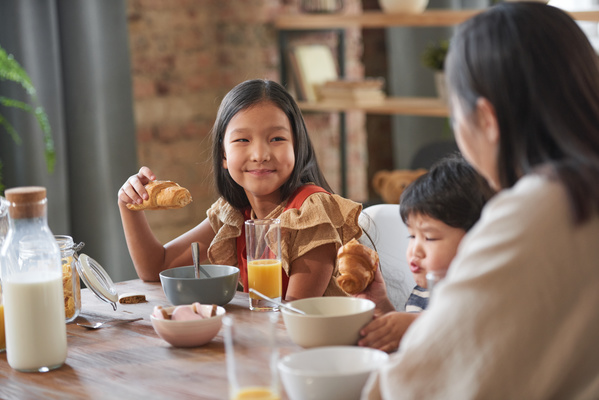 A dark-haired girl with a croissant in her hand smiles sitting at the breakfast table with her mother and younger brother