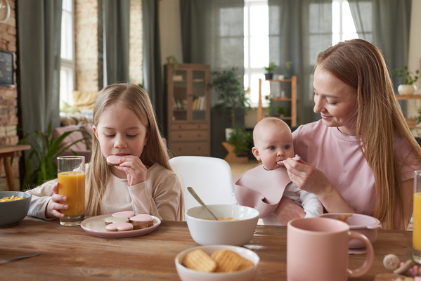 A girl with blonde long hair eats gingerbread in icing for breakfast sitting at the table with her mother feeding the baby in a bib