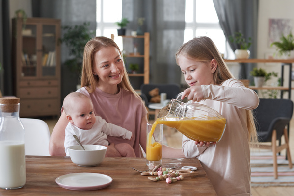A blonde girl pours orange juice from a jug into a glass to a smiling mother with a baby in her arms