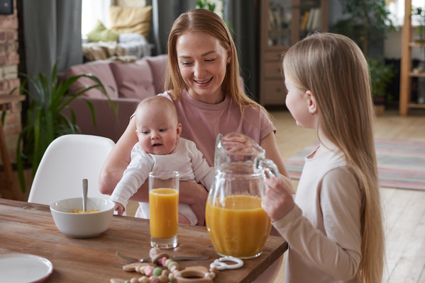 A long-haired smiling mother with a baby in her arms is sitting at the breakfast table next to her daughter holding the handle of a jug of orange juice
