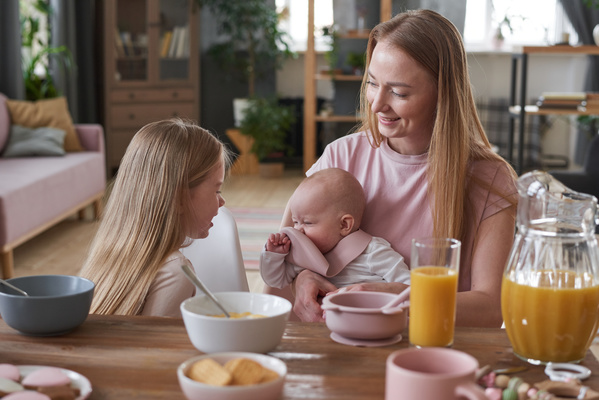 A long-haired smiling mother with a baby playing with a bib sits at the breakfast table next to her daughter looking at the baby