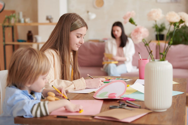 A teenage girl sitting at a table with her younger brother draw illustrations on homemade postcards dedicated to Mothers Day