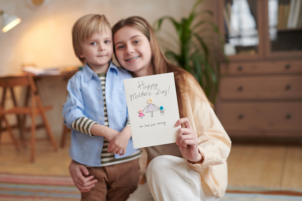 A girl with long blonde hair holds a handmade Mothers Day card with button applique hugging her younger brother
