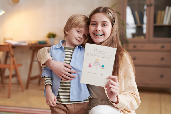 A girl with long blonde hair with a younger brother smiling holds a homemade mothers Day card with a button applique