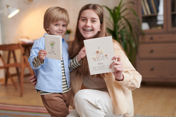 A little boy with a long-haired sister squatting hold homemade Mothers Day cards with drawings and button applications