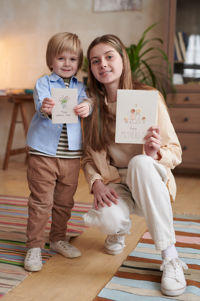 A little boy and a long-haired sister squatting with handmade Mothers Day cards with drawings and button applications in hands