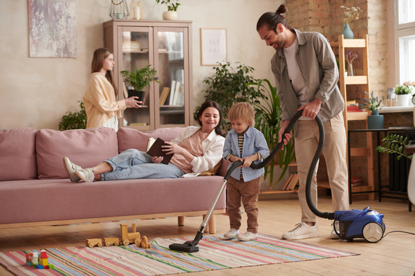 A woman with a book lying on the sofa looks at her husband and young son vacuuming the carpet on Mothers Day
