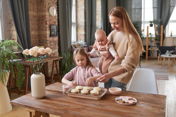 A mother with long hair holding a baby in her arms decorate cupcakes with confectionery sprinkles with her eldest daughter