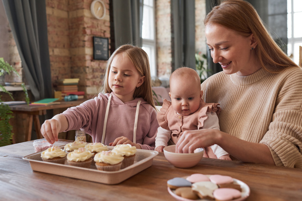 A mother with long hair with a baby in her arms and her eldest daughter sitting at a table sprinkle cupcakes with white cream