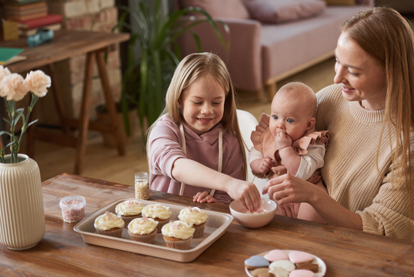 A girl takes a pastry sprinkle for cupcakes sitting with her mother holding the baby in her arms at a table on which there are cakes