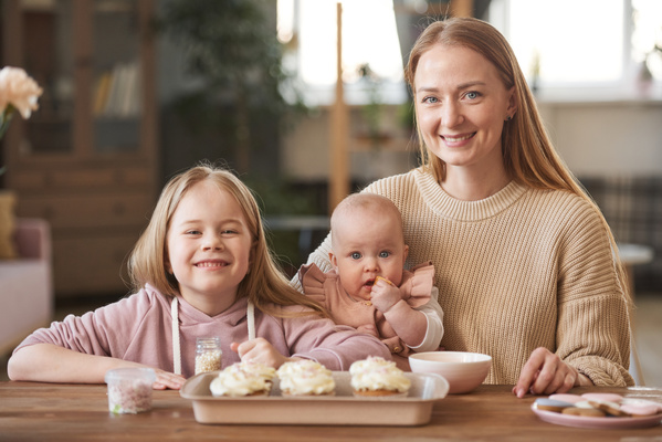 A mother with a baby in her arms and a smiling eldest daughter sitting at a table with cupcakes on a tray
