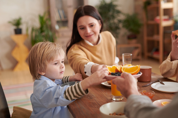 A dark-haired mother smiling and passing a plate of oranges to her husband and young son during family breakfast