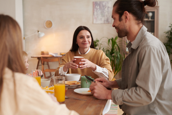 A dark-haired mother with a brown mug in her hands looks tenderly at her husband sitting with him and the children at the table during family breakfast