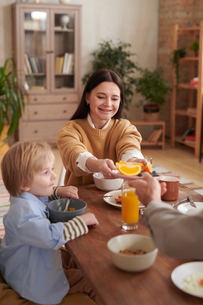 A dark-haired mother smiling passes a plate of oranges to her husband while sitting at the table with him and her young son at a family breakfast